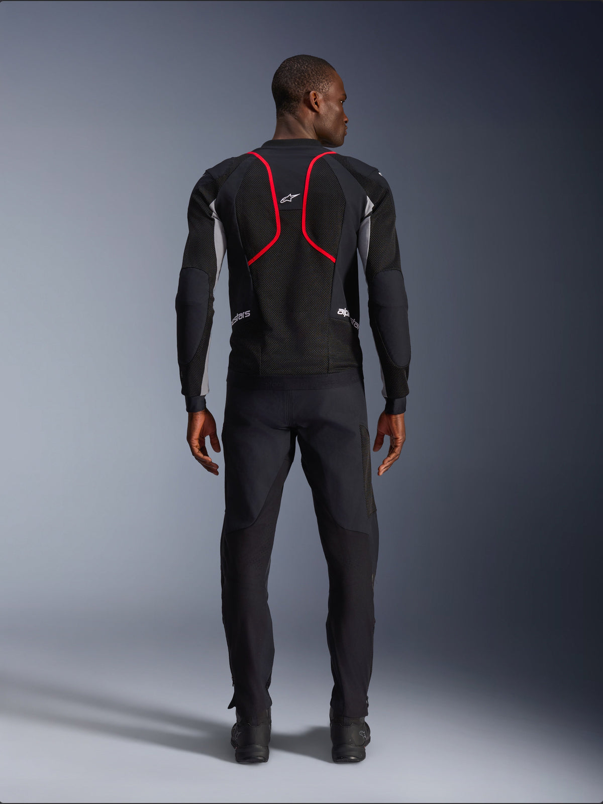 Modelo visto de costas a usar o Casaco Alpinestars Troop-Air Pro, preto com detalhes em cinza cinza e vermelho vivo, apresentando construção em malha técnica com debrum vermelho proeminente nas costas, logótipo Alpinestars na parte superior das costas, combinado com calças de motociclismo pretas, casaco de ar blindado premium para condução em estrada