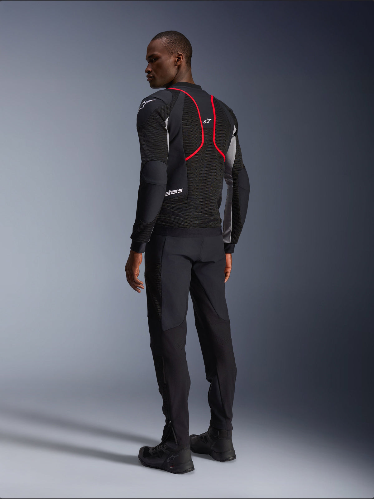 Alpinestars Troop-Air Pro Jacket, casaco de motociclismo, preto, cinzento e vermelho vivo, vista de costas num modelo, com linhas de realce vermelhas e painéis de ventilação em malha, usado com calças e botas pretas, estilo de motociclismo urbano