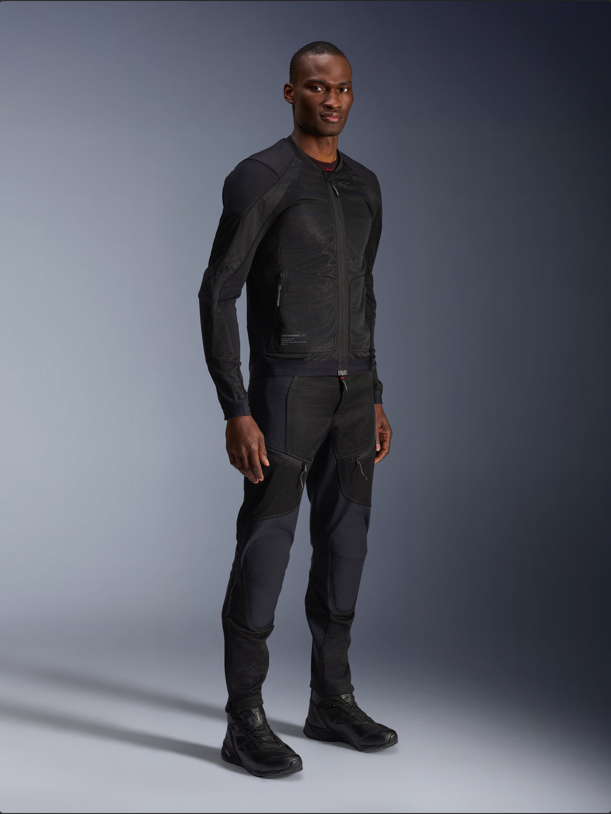Alpinestars Troop-Air Jacket, casaco de motociclismo em malha técnica, preto, usado por um modelo com calças e botas pretas a condizer, com construção em malha leve e um design urbano minimalista