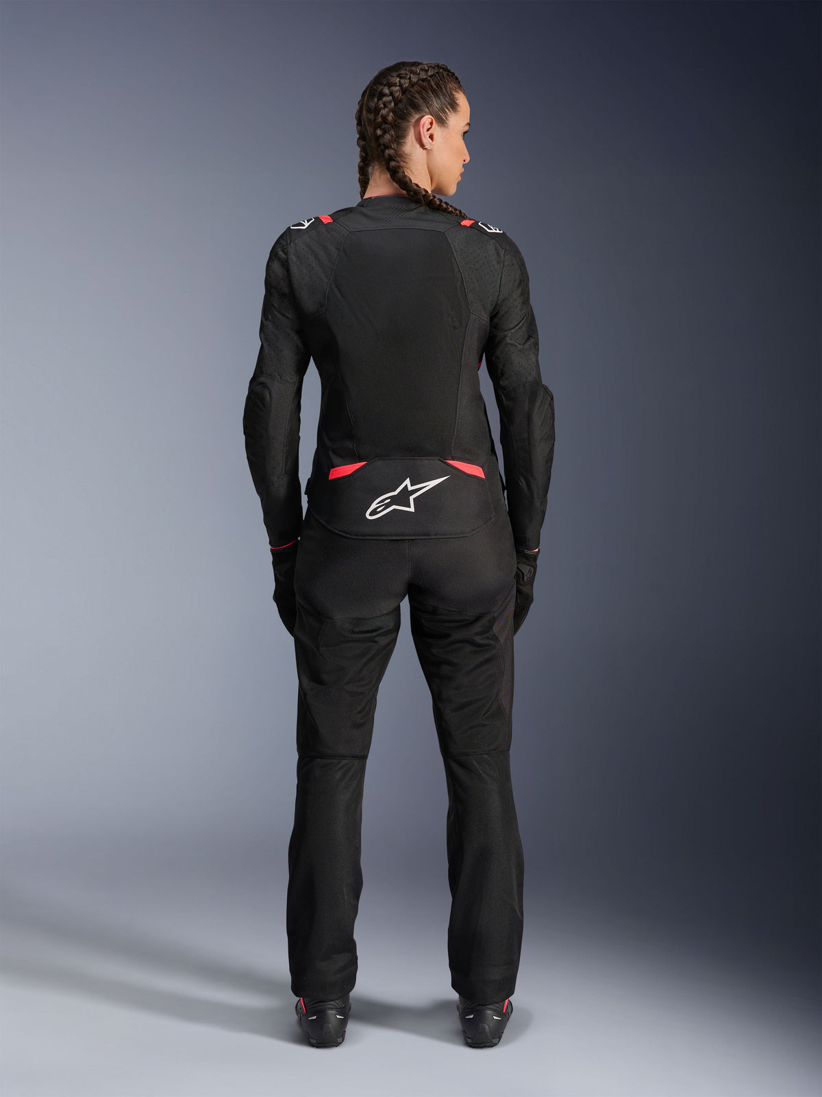 Casaco Alpinestars Stella T-SPS Air V2, casaco de motociclismo têxtil para mulher, preto e Diva Pink, vista traseira numa modelo com cabelo entrançado, com grandes painéis de malha para ventilação, logótipo Astars branco na parte inferior das costas, combinado com calças de condução pretas a condizer.