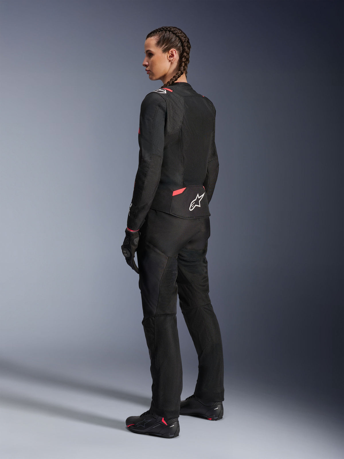 Alpinestars Stella T-Sps Air v2 Jacket para Mulher, preto e Diva Pink, casaco de motociclismo em têxtil visto de uma perspetiva lateral traseira, com grandes painéis de malha para circulação de ar e logótipo de estrela branco nas costas, usado com calças, luvas e botas de motociclismo pretas a condizer.