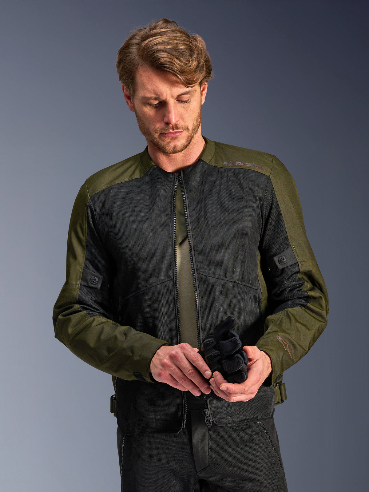 Homem a usar o Casaco Alpinestars C-1 Air em Khaki, casaco de motociclismo urbano com tecido verde azeitona e painéis de malha pretos, a olhar para baixo enquanto segura luvas de motociclismo pretas, estilo limpo e contemporâneo para condução em estrada.