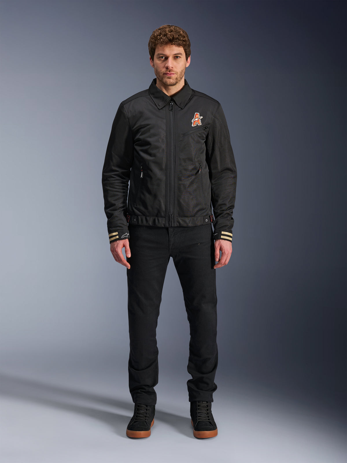 Modelo masculino a usar o Casaco Alpinestars Ignite Air em preto, com gola de camisa clássica, fecho de correr frontal e logótipo cor de laranja no peito. Combinado com calças pretas e sapatilhas pretas de cano alto para um visual de motociclismo urbano contemporâneo.