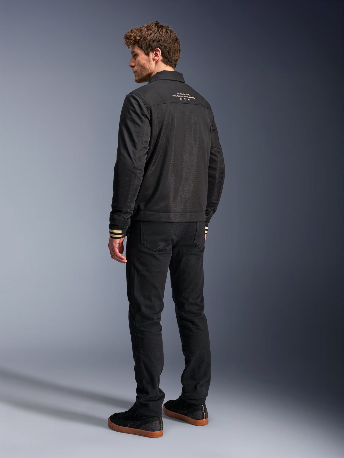 Casaco Alpinestars Ignite Air, casaco de motociclismo preto, vista traseira com texto gráfico branco na parte superior das costas e punhos com riscas bege, silhueta de estilo Harrington com corpo em malha, combinado com calças pretas e sapatilhas pretas