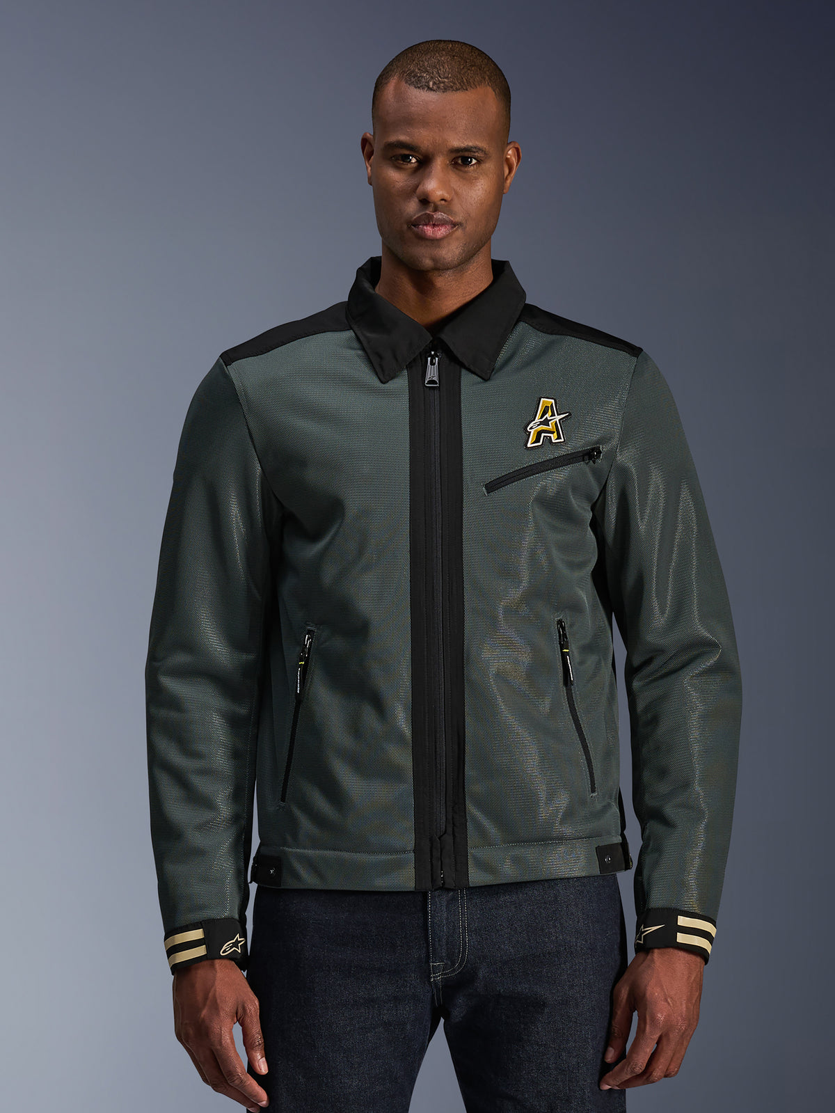Alpinestars Ignite Air Jacket, casaco de motociclismo urbano, verde floresta escuro com detalhes pretos, apresentado num modelo com calças de ganga, gola estilo Harrington, fecho de correr frontal, logótipo no peito e punhos com riscas, construção moderna em malha e têxtil para o verão