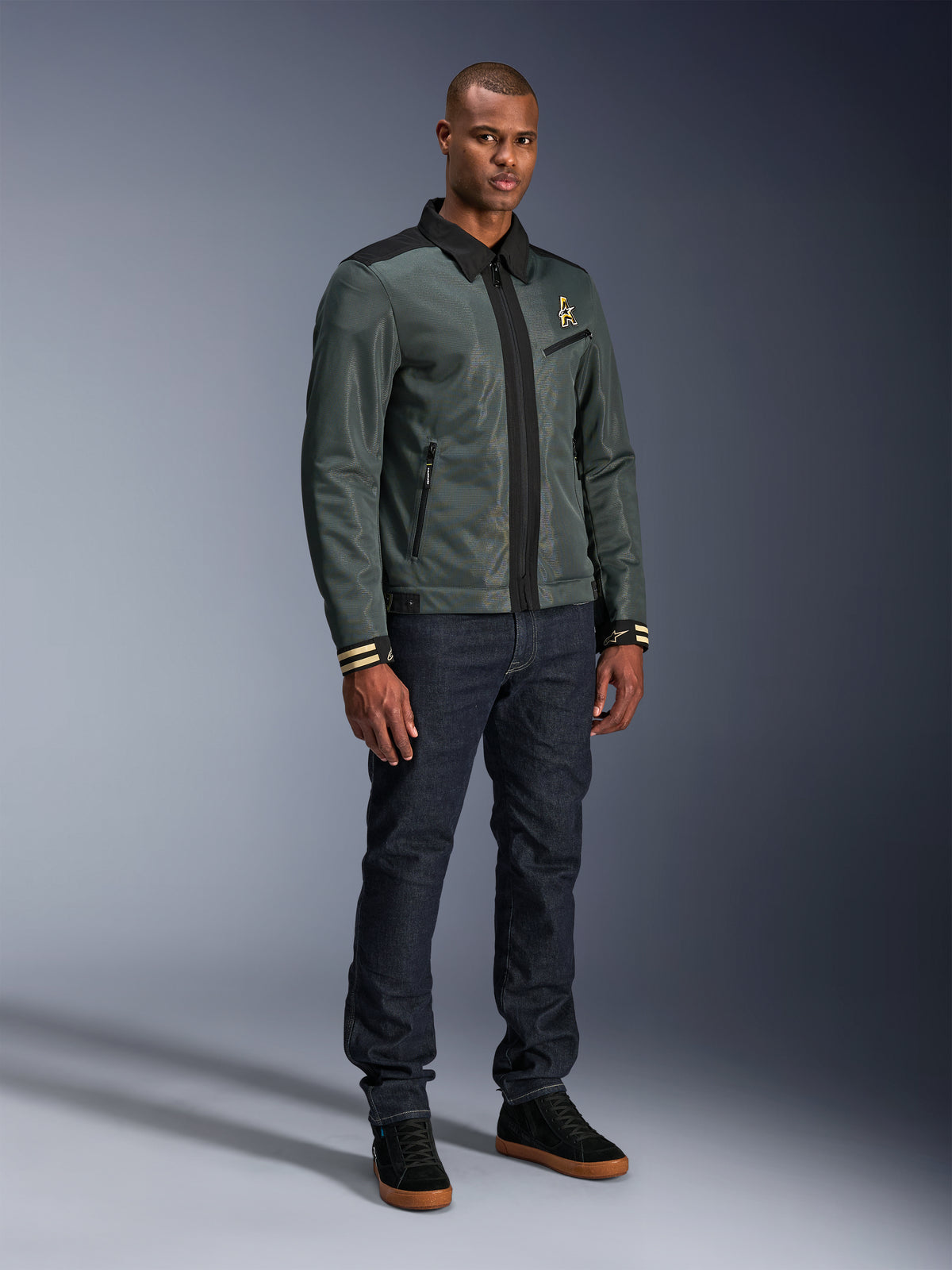 Casaco Alpinestars Ignite Air, Urban Gray Black Blue, casaco de motociclismo urbano, usado por um modelo com calças de ganga escuras e ténis pretos, apresentando corpo em malha, gola de camisa tradicional, silhueta inspirada no estilo Harrington e logótipo Alpinestars em dourado.