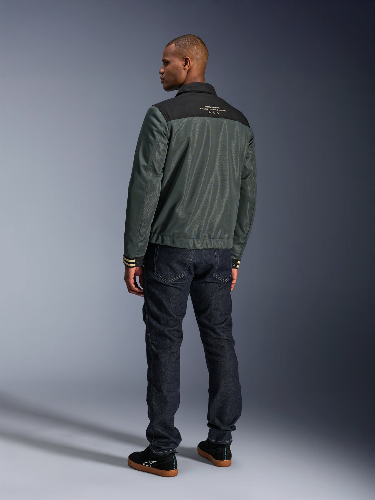 Casaco Ignite Air da Alpinestars em Cinza Urbano, Preto e Azul, vista traseira no modelo, com corpo em malha, escapulário preto e texto branco nos ombros, punhos às riscas, combinado com calças de ganga escuras e sapatilhas pretas, vestuário de motociclismo de estilo urbano