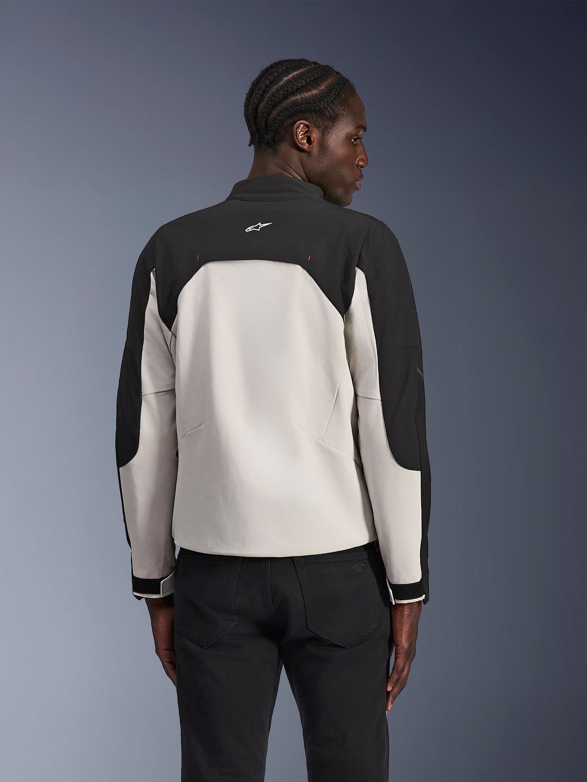 Alpinestars Provoke Jacket, casaco de motociclismo urbano, Black Metal Gray, vista traseira num modelo, com um design têxtil bicolor com ombros pretos e painel traseiro cinzento claro, logótipo de estrela Alpinestars na parte de trás da gola, combinado com calças pretas sobre um fundo em degradê azul