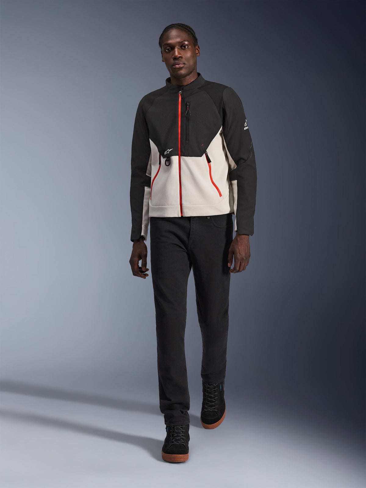 Modelo masculino a usar o Casaco Alpinestars Provoke, casaco de motociclismo urbano em preto e cinzento metalizado com pormenores de fechos vermelhos, combinado com calças pretas e sapatilhas pretas de cano alto, equipamento de proteção moderno de estilo urbano para condução na cidade