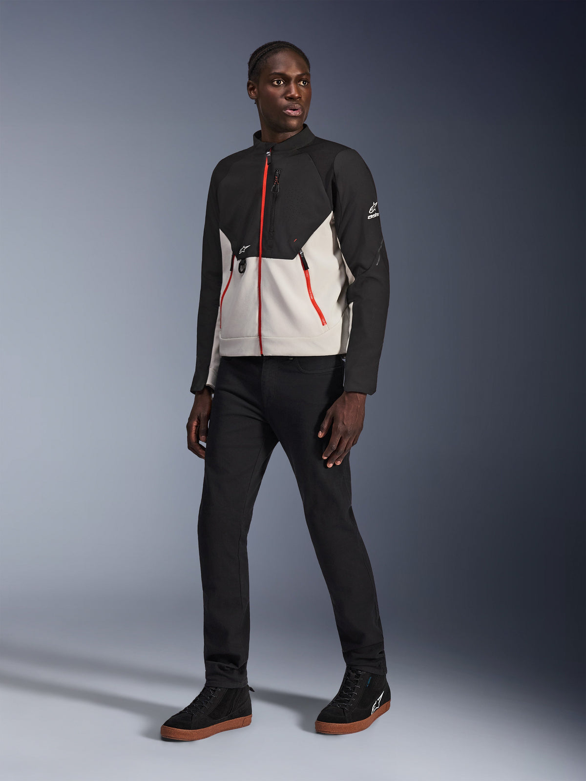 Modelo a usar o Casaco Alpinestars Provoke em Black Metal Gray, com fechos frontais e de bolsos em vermelho, combinado com calças pretas e sapatilhas de cano alto pretas com sola de cor mel; equipamento de motociclismo urbano contemporâneo para um visual citadino.
