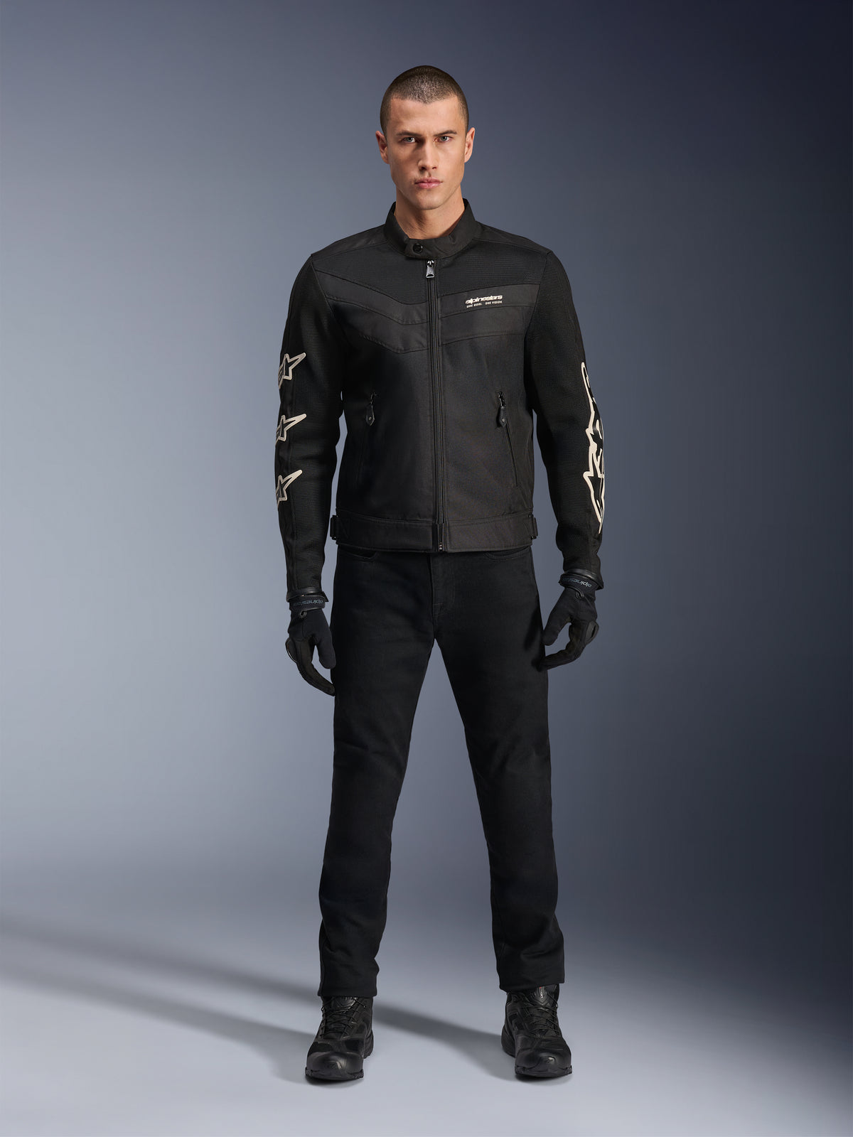 Modelo masculino a usar o Casaco Alpinestars T-Dyno Air, preto, casaco de motociclismo têxtil leve com malha 3D respirável, logótipos de estrela brancos nas mangas, combinado com calças, luvas e botas pretas, vista frontal de pé