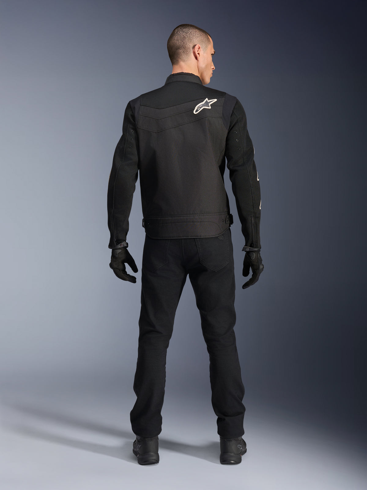 Casaco Alpinestars T-Dyno Air, preto, vista traseira no modelo, casaco de motociclismo têxtil com painéis de malha 3D e logótipo de estrela branco no ombro, usado com calças e luvas pretas, visual de condução urbana moderno
