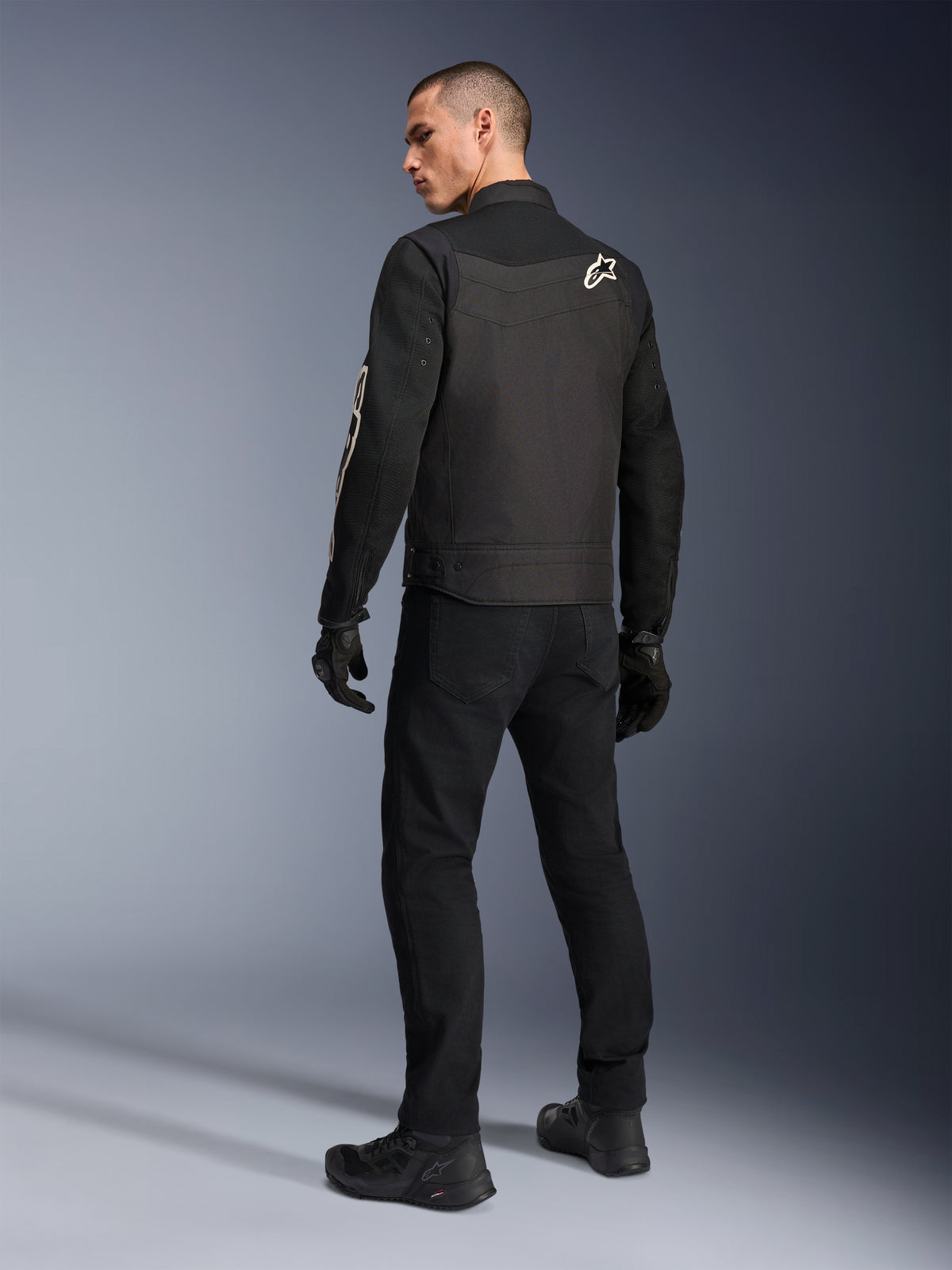 Alpinestars T-Dyno Air Jacket, preto, vista traseira de modelo a usar um casaco de motociclismo têxtil respirável, com logótipo Alpinestars branco no ombro, combinado com calças e luvas pretas, estilo de condução urbana