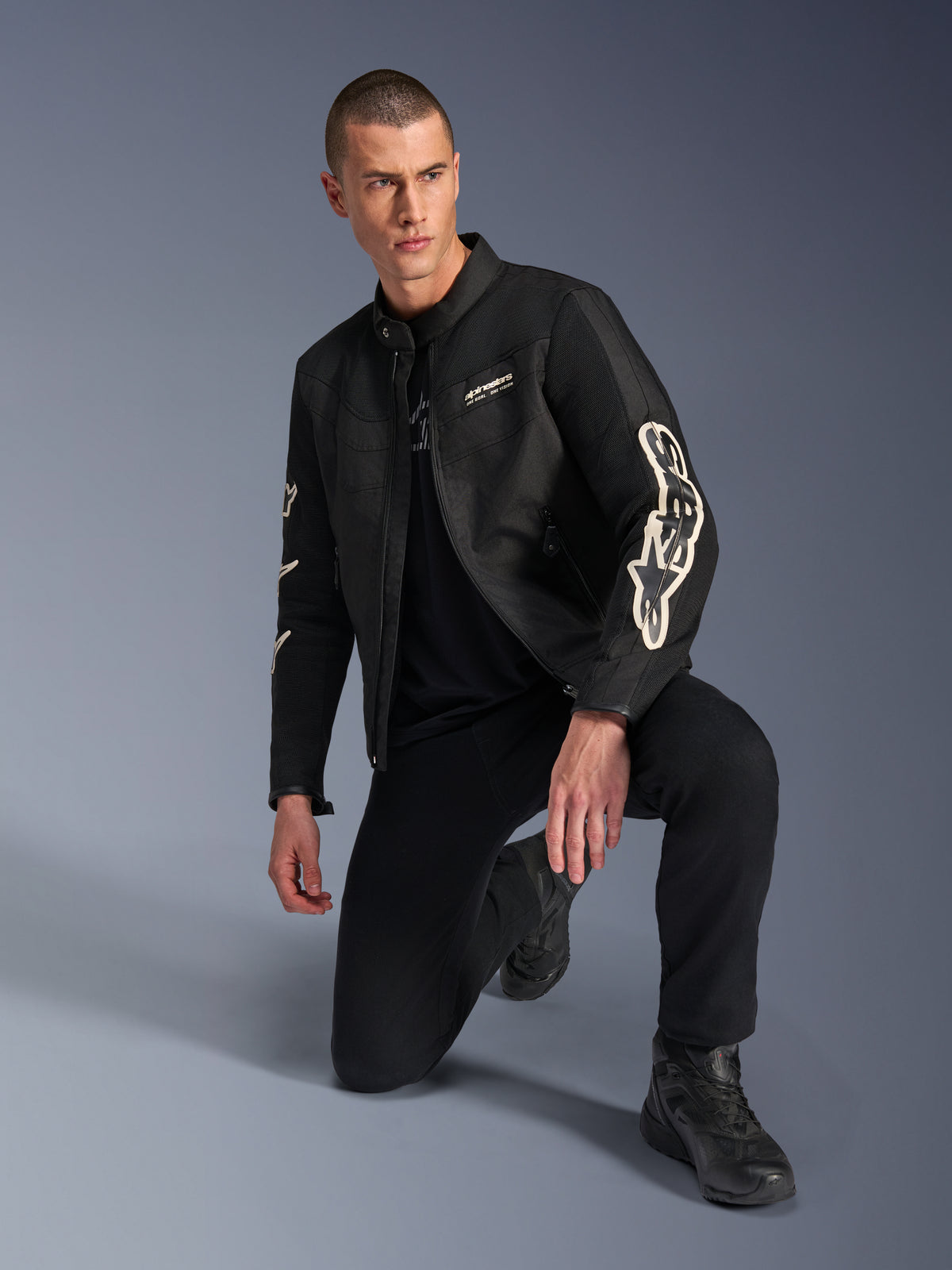 Modelo masculino a usar o Casaco Alpinestars T-Dyno Air, casaco de motociclismo têxtil preto com logótipos brancos grandes nas mangas, combinado com calças pretas e botas de motociclismo, design moderno de inspiração vintage com painéis de malha 3D.