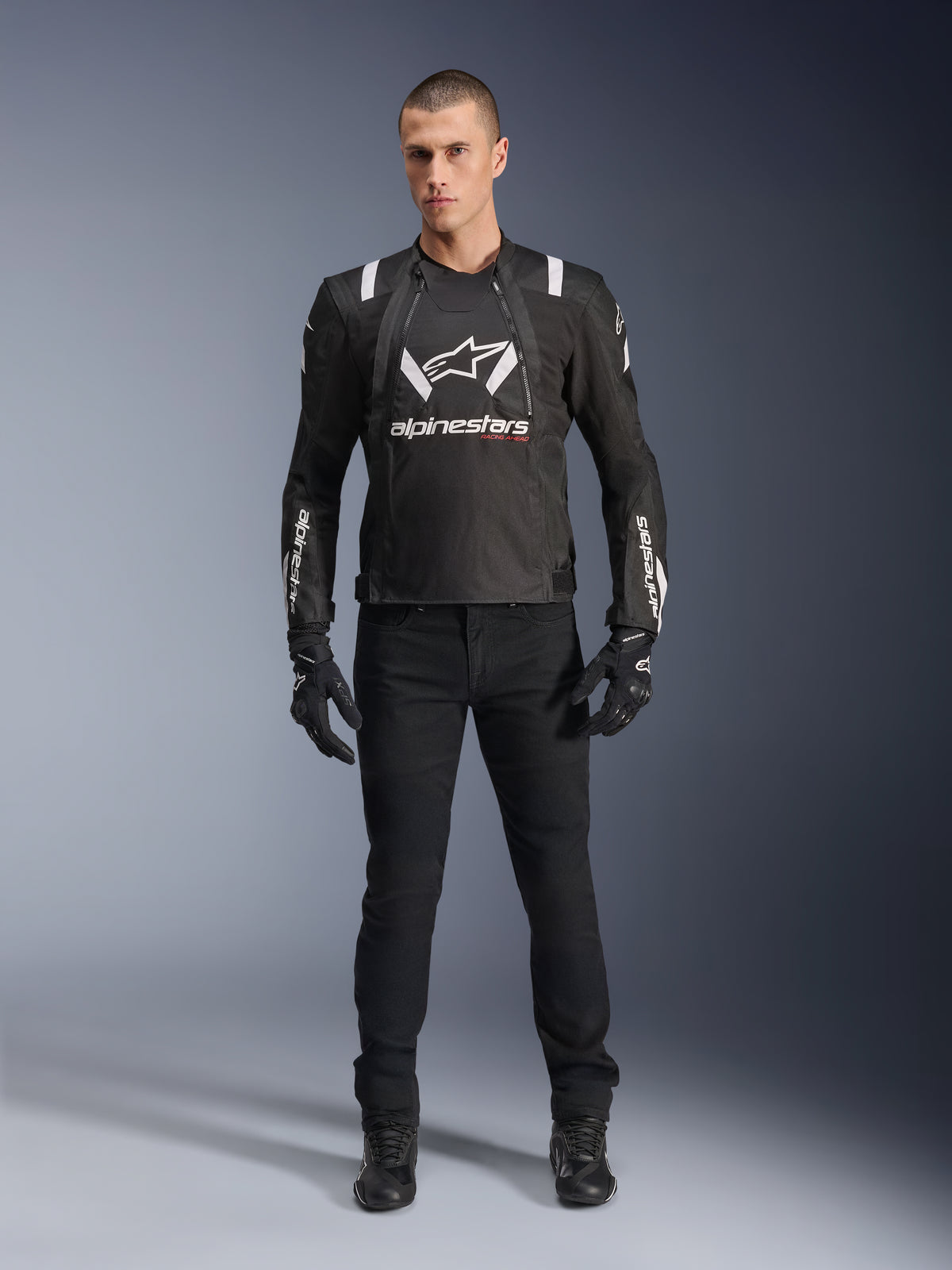 Casaco Alpinestars T-Stunt Air, casaco de motociclismo preto e branco, usado com calças de condução, luvas e botas pretas, com logótipo Alpinestars em destaque a branco no peito e nas mangas, design têxtil de corte desportivo com painéis em malha para ventilação