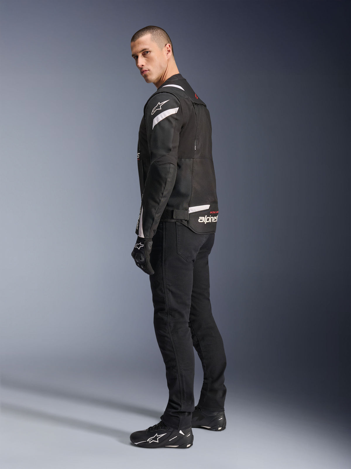 Alpinestars T-Stunt Air Jacket, casaco modular de motociclismo em preto e branco com painéis de malha técnica, visto de perfil lateral traseiro num modelo, usado com calças e sapatos de motociclismo pretos, com detalhes e logótipo em branco nos ombros e costas