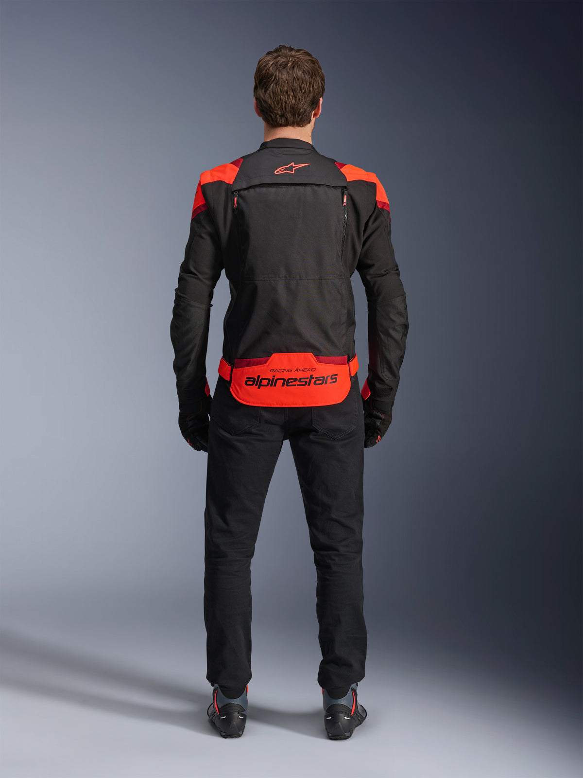 Casaco Alpinestars T-Stunt Air, preto com detalhes em vermelho fluo e vermelho escuro, vista traseira no modelo, combinado com calças e botas de condução pretas, com logótipo Alpinestars proeminente no painel inferior traseiro, design modular com construção em malha para ventilação.
