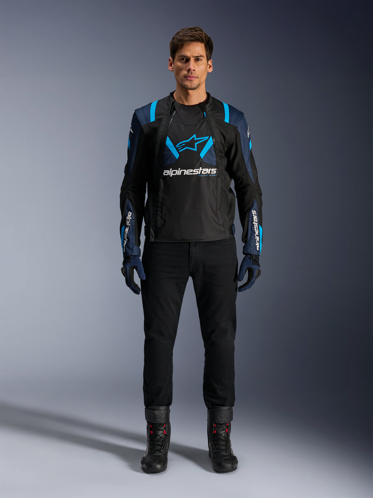 Alpinestars T-Stunt Air Jacket, casaco de motociclismo, preto com detalhes em azul escuro e azul vivo, logótipo branco grande no peito, construção em malha com corte desportivo para ventilação, usado com calças e botas de condução pretas para uma estética de condução em estrada elegante