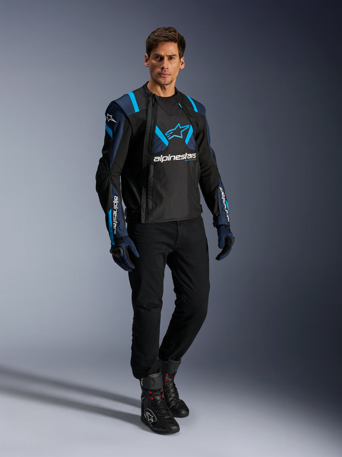 Modelo masculino a usar o casaco Alpinestars T-Stunt Air, em preto, azul escuro e azul brilhante, com painéis de malha proeminentes para fluxo de ar, combinado com calças pretas e luvas de condução; casaco de motociclismo com corte desportivo para condução em estrada no verão.