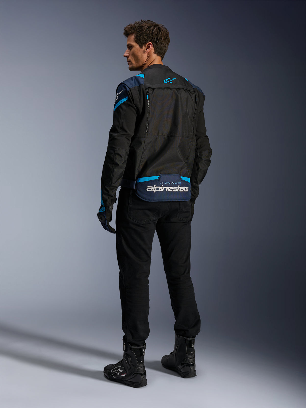 Casaco Alpinestars T-Stunt Air, casaco de motociclismo, preto com detalhes em azul escuro e azul Blithe, vista traseira mostrando painéis de malha modulares e o logótipo na zona lombar, usado por um modelo com calças e botas de motociclismo pretas.