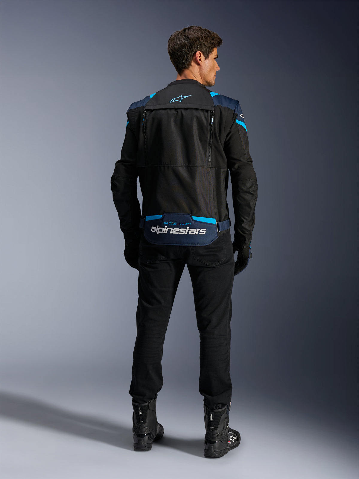 Casaco Alpinestars T-Stunt Air, casaco modular de motociclismo em têxtil, preto, azul escuro e azul vibrante, vista traseira que mostra o compartimento de arrumação na zona lombar com logótipo Alpinestars a branco, usado com calças e botas pretas.