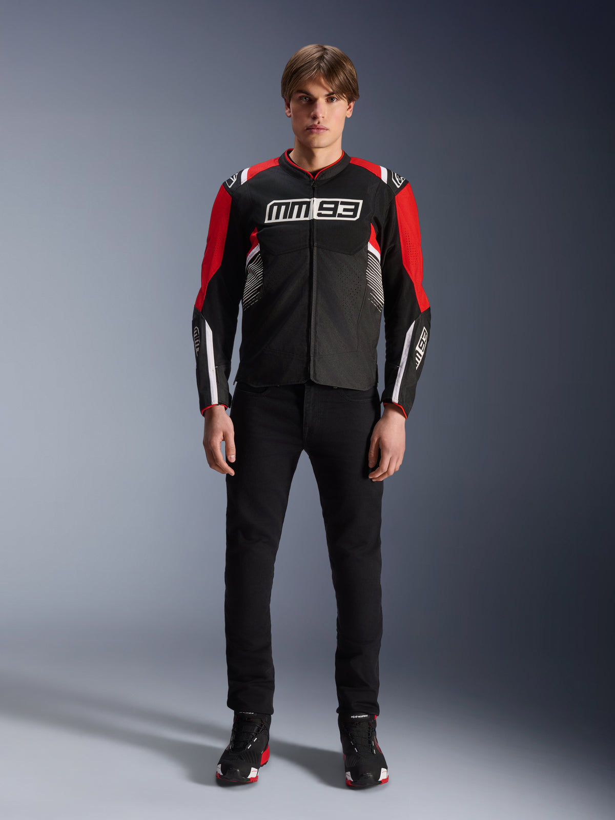Male model wearing the Alpinestars MM93 T-SPS Air V2 Jacket in Black, Bright Red, and White, featuring Marc Márquez MM93 branding on the chest, mesh paneling for airflow, paired with black pants and sneakers in a studio setting.