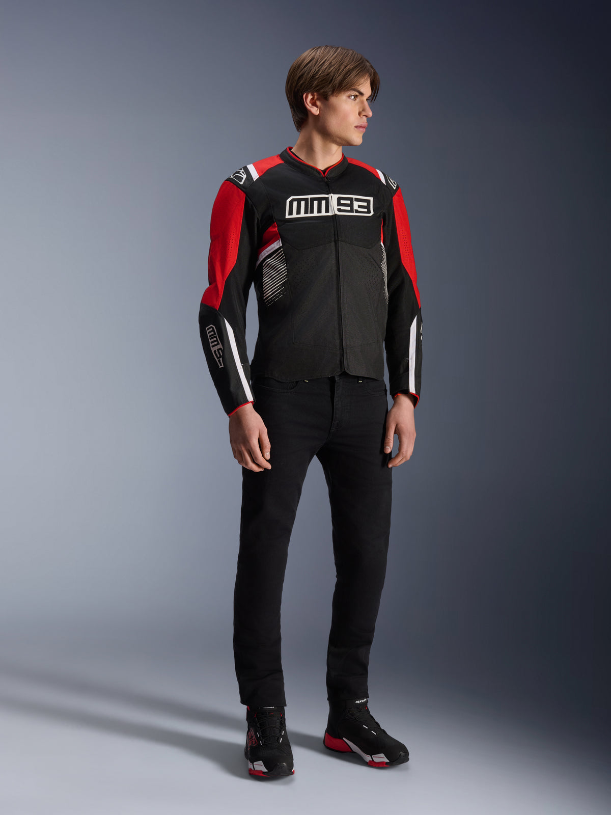 Male model wearing the Alpinestars MM93 T-SPS Air v2 Jacket in Black Bright Red White, featuring prominent MM93 branding on the chest and sleeves, paired with black pants and riding boots, sporty urban motorcycle style with visible mesh panels for airflow.