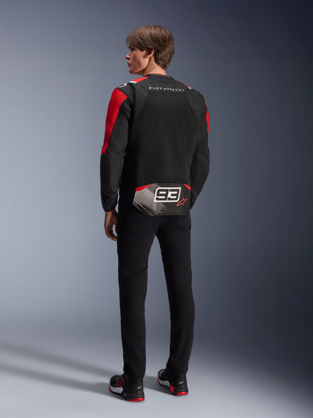 Model from behind wearing the Alpinestars MM93 T-SPS Air V2 Jacket, Black with Bright Red and White details, featuring 'MARC MARQUEZ' text and large '93' logo on the back tail, mesh textile construction for ventilation, worn with black pants and coordinated black and red riding shoes