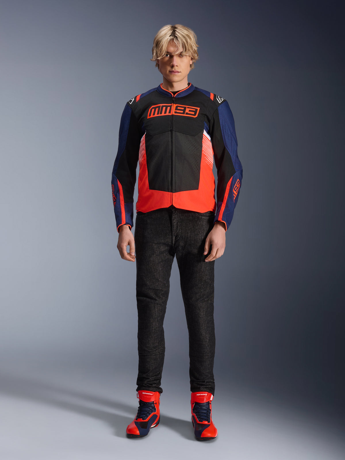 Casaco Alpinestars MM93 T-SPS Air V2, azul-marinho e preto com detalhes em vermelho fluo, com o logótipo MM93 no peito, usado com calças de motociclismo pretas e botas azuis e vermelhas a condizer, equipamento de motociclismo urbano