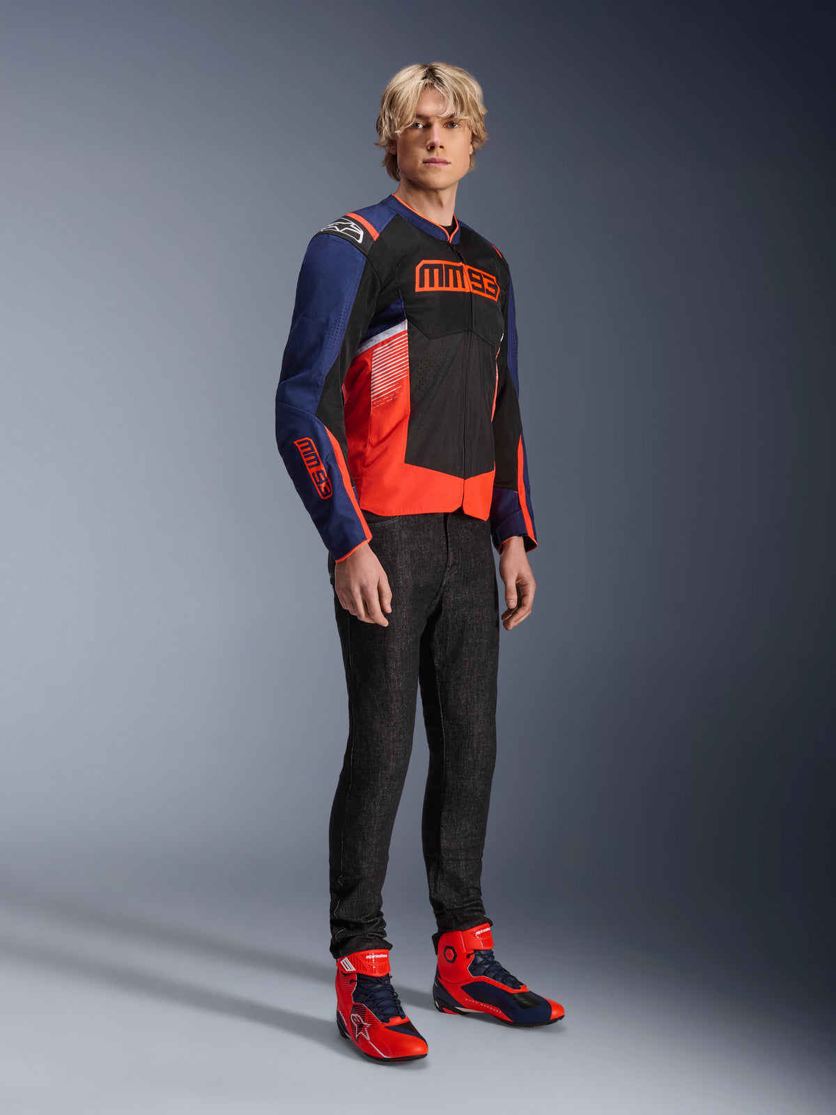 Modelo masculino a usar o casaco Alpinestars MM93 T-SPS Air V2 em azul marinho, preto e vermelho fluo, com o logótipo MM93 em destaque no peito e nas mangas, combinado com calças de ganga escuras e botas de motociclismo vermelhas e azuis a combinar, equipamento de condução urbana desportivo