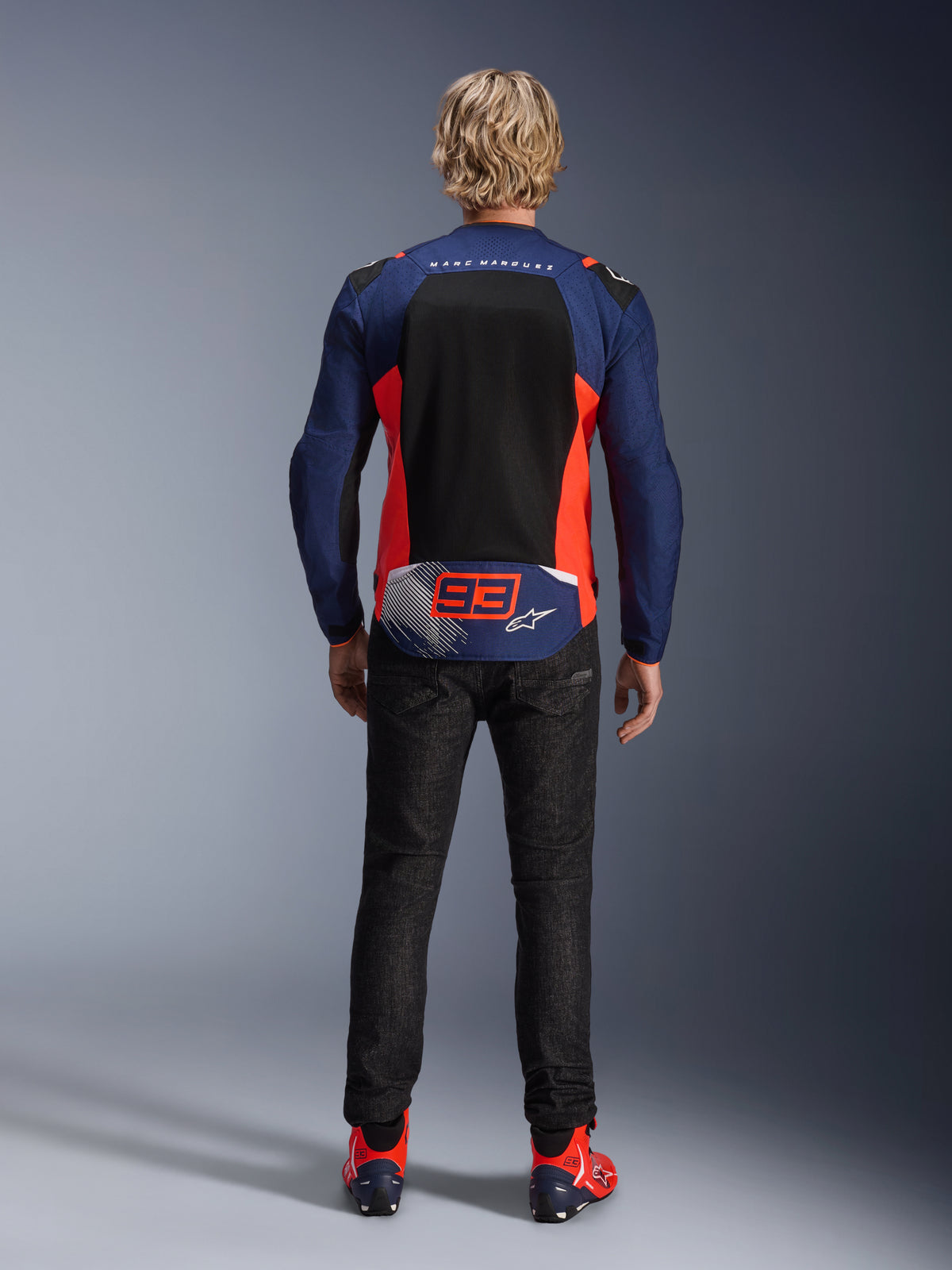 Vista posterior de um modelo a usar o Casaco MM93 T-SPS Air V2 em azul marinho com pormenores em vermelho fluo e preto, com um grande painel de malha nas costas e a marca '93' de Marc Marquez, combinado com calças de ganga escura e botas de motociclismo a condizer