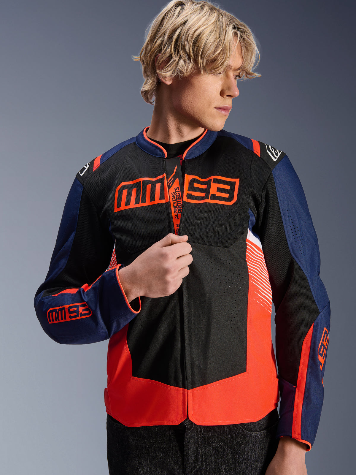 Casaco Alpinestars MM93 T-SPS Air V2, casaco de motociclismo têxtil em azul-marinho, preto e vermelho fluorescente, vestido por um modelo masculino a ajustar o fecho, com o logótipo MM93 de Marc Márquez no peito e nas mangas, e painéis de malha ventilada para condução em estrada no verão.