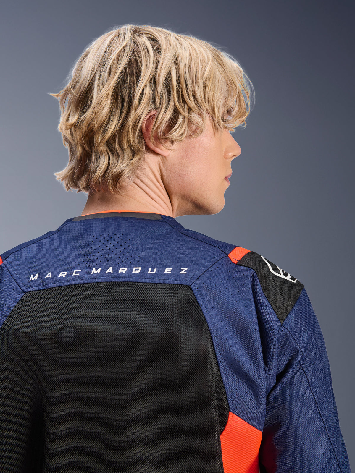 Casaco Alpinestars MM93 T-SPS Air V2, azul-marinho e preto com pormenores em vermelho fluo, grande plano da parte superior das costas com a marca 'MARC MARQUEZ', painéis de malha respirável e construção têxtil perfurada, vestido por um modelo loiro.