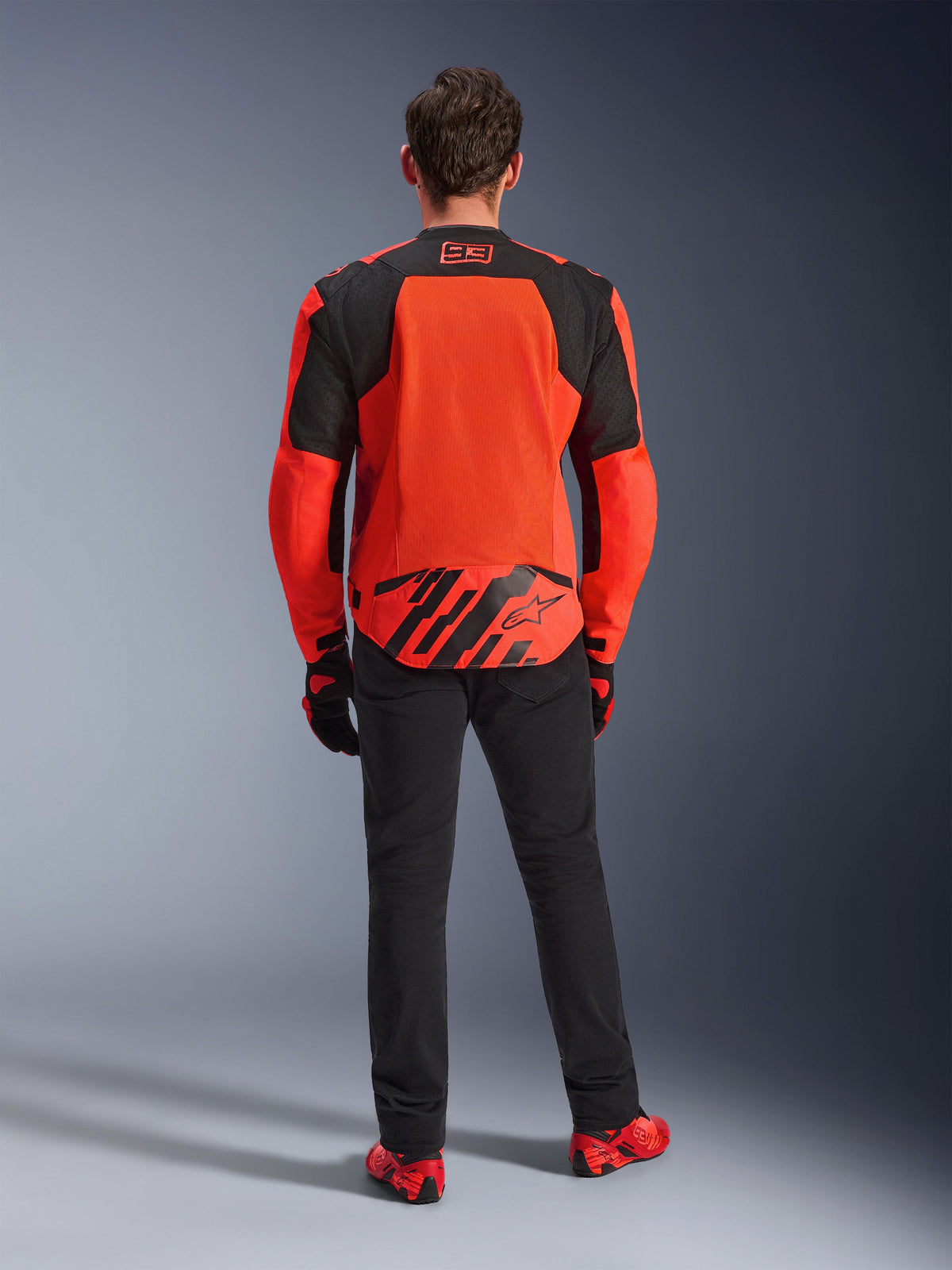 Vista traseira do Casaco Alpinestars MM93 T-SPS Air V2, vermelho fluorescente e preto, casaco têxtil de motociclismo com grandes painéis de malha nas costas para ventilação, usado com calças pretas e calçado vermelho, apresentando a marca Marc Marquez 93