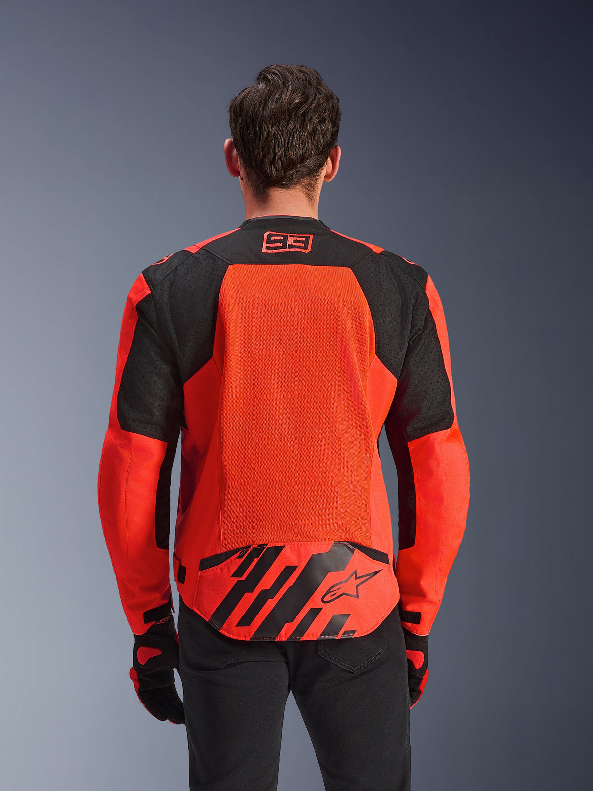 Casaco Alpinestars MM93 T-SPS Air V2, vista traseira, vermelho fluo e preto, com logótipo de corrida 93 na gola, grande painel de malha nas costas para ventilação, aba lombar com o logótipo da estrela Alpinestars e riscas geométricas, usado com calças pretas e luvas de motociclismo.