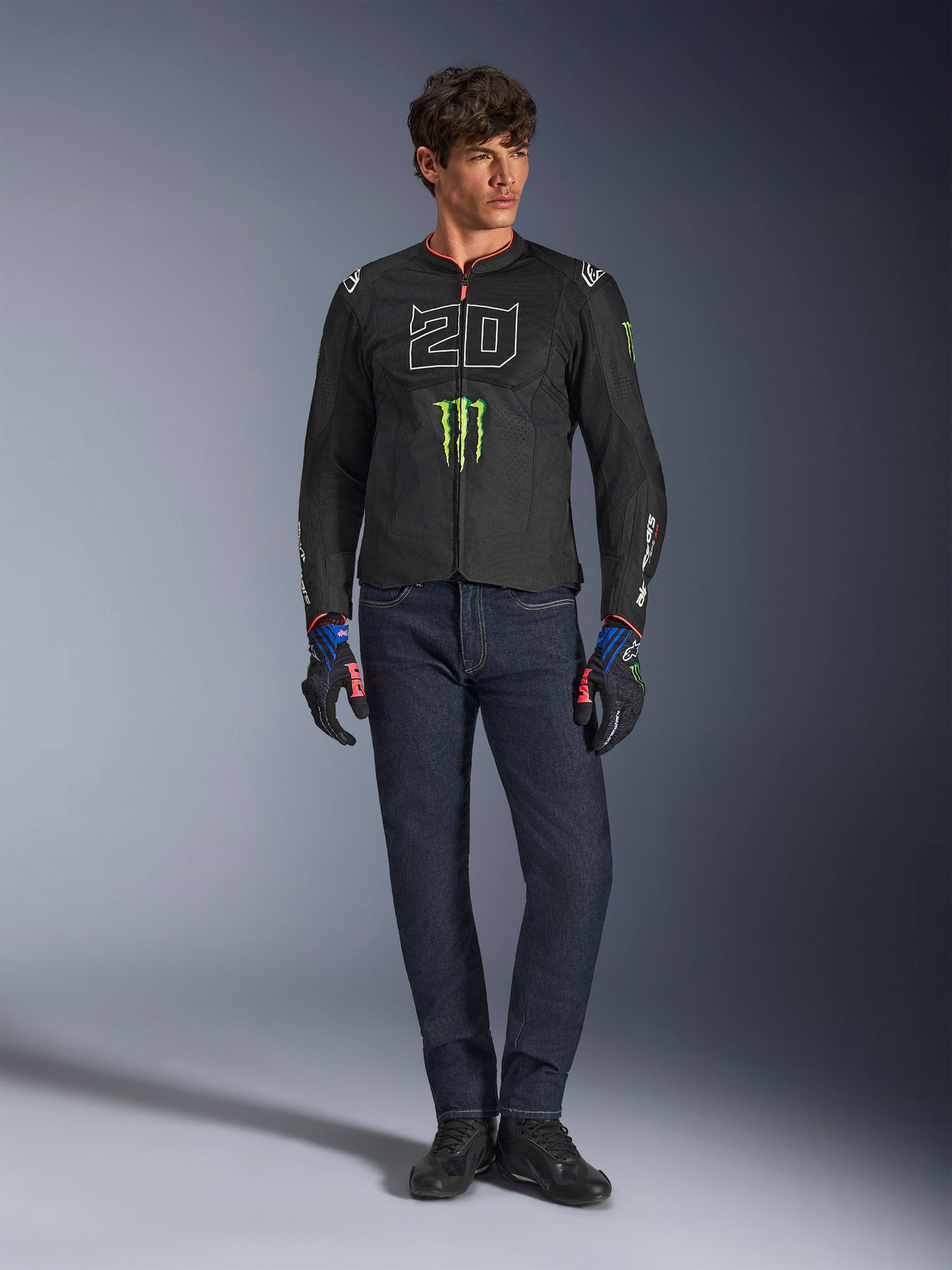 Piloto com o Casaco Alpinestars FQ20 T-SPS Air V2 Monster, preto com logótipo verde Monster Energy e gráfico '20' branco no peito, combinado com calças de ganga azul escuro e luvas técnicas de condução, equipamento de motociclismo desportivo urbano.