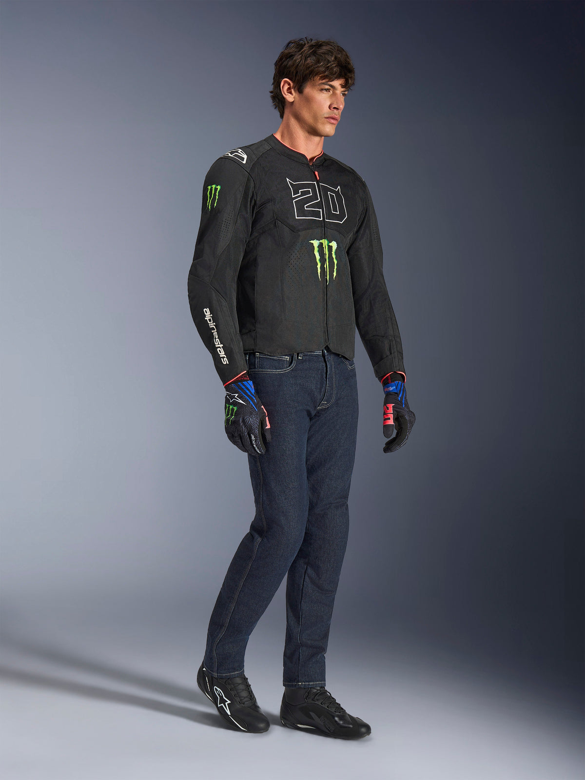 Motociclista a usar o casaco Alpinestars FQ20 T-SPS Air V2 Monster em preto, com o logótipo '20' e a garra da Monster Energy no peito, combinado com calças de ganga de motociclismo escuras, sapatos de motociclismo pretos e luvas FQ20 a combinar, fotografia de estúdio de corpo inteiro.