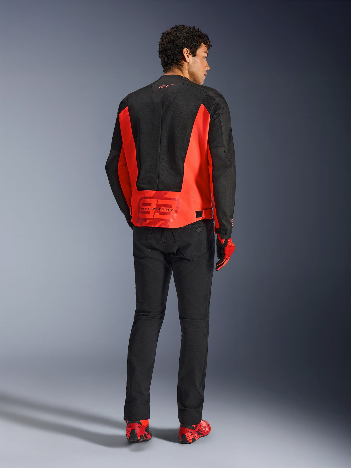 Rear view of a male model wearing the Alpinestars MM93 C-1 Air Jacket in Red Fluo and Black, featuring the Marc Marquez 93 logo on the lower back, paired with black pants and red riding shoes, urban motorcycle commuting style