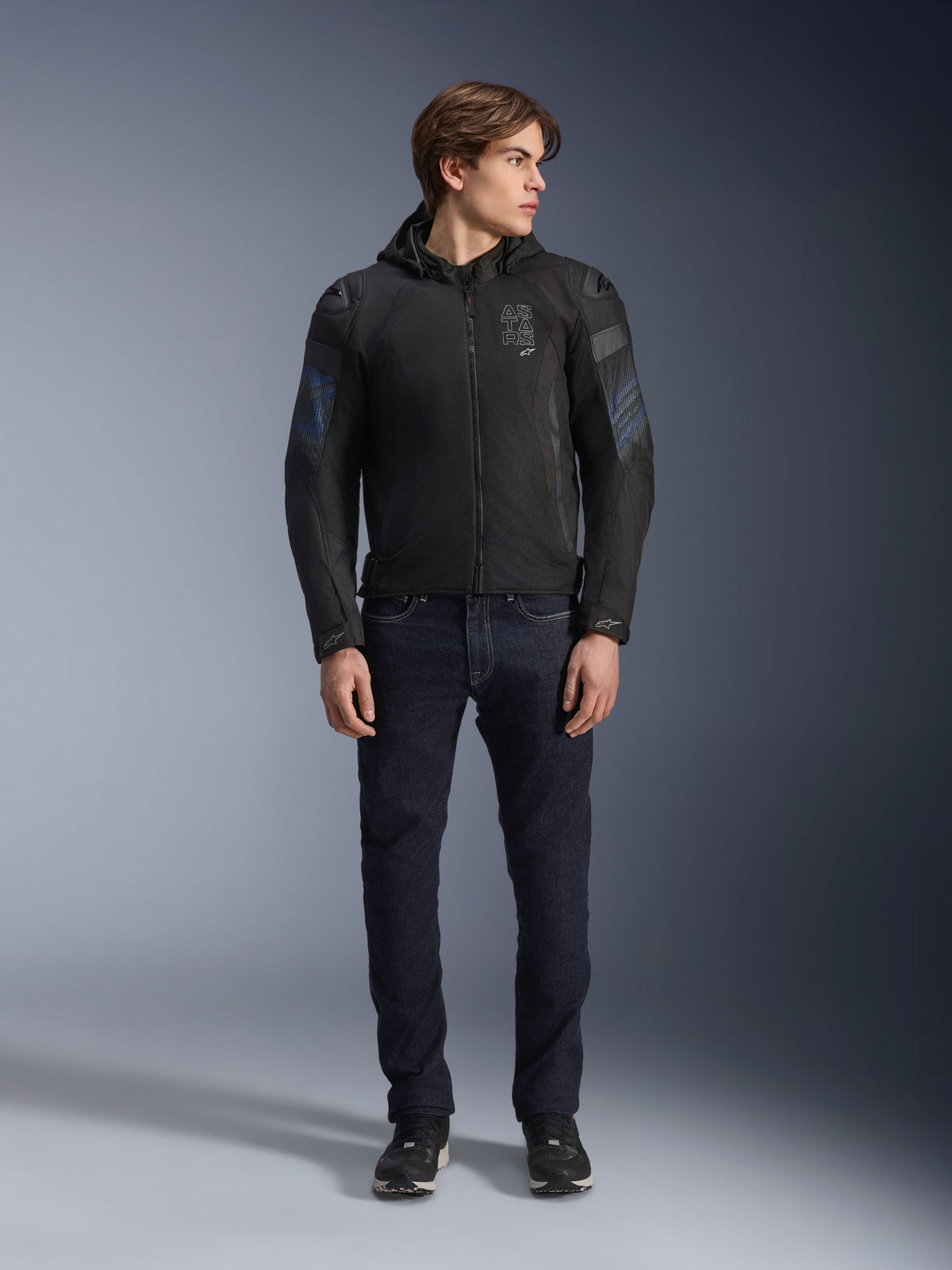Male model wearing Alpinestars Zaca Air Venom WP Jacket in Black and Electric Blue, textile motorcycle jacket with 3D mesh panels and a detachable hood, paired with dark denim jeans, modern sport-inspired riding gear