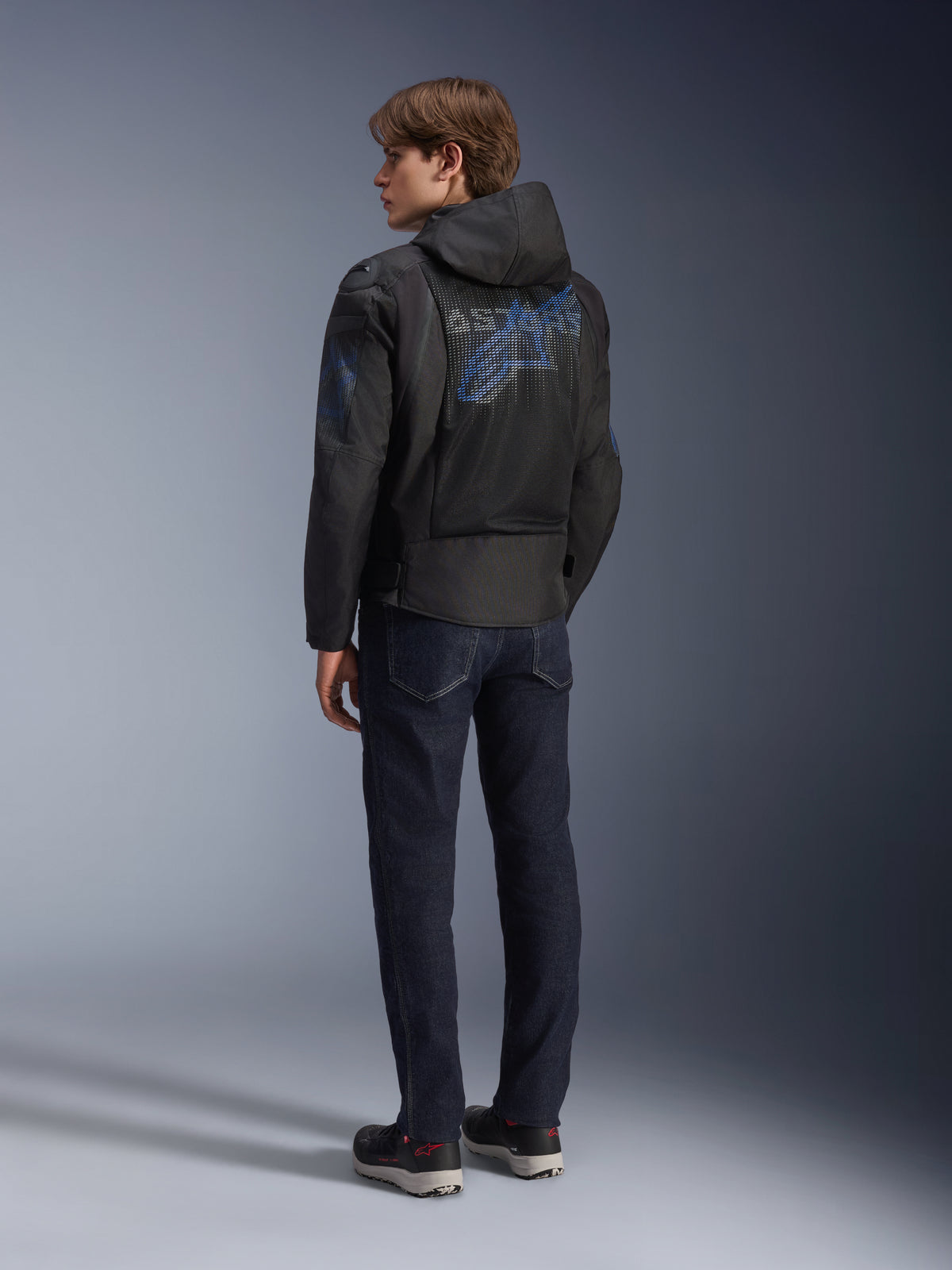 Alpinestars Zaca Air Venom WP Jacket, Black and Electric Blue, rear view on a model, mesh textile motorcycle jacket with blue Astar logo on the back, featuring a detachable hood, worn with dark jeans and black riding shoes.