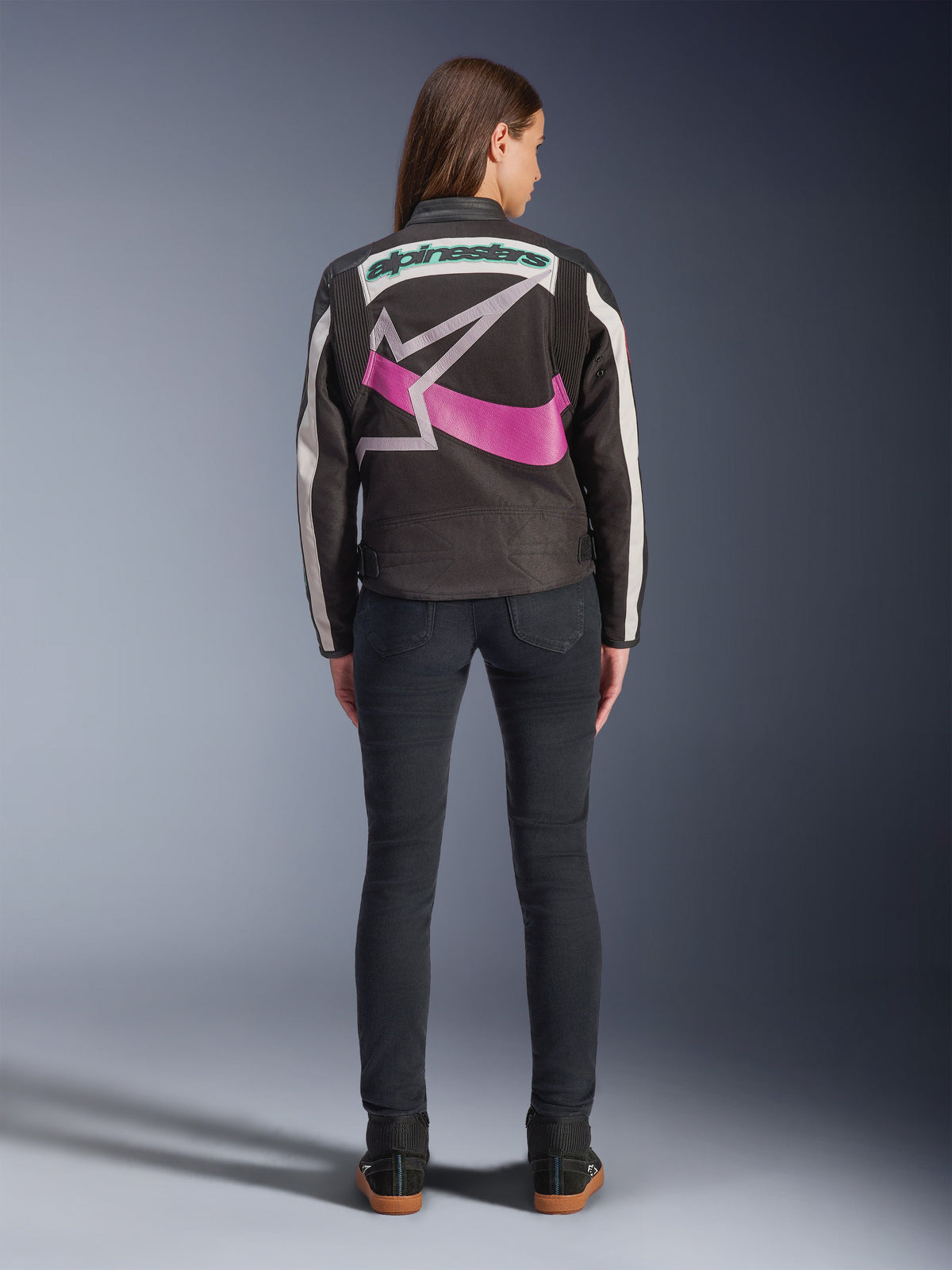 Vista traseira de uma mulher a usar o Casaco Alpinestars Idles, preto com riscas off-white, com um grande logótipo de estrela da Alpinestars em roxo e branco e marca nominativa verde nas costas, combinado com calças de ganga slim-fit escuras, vestuário de motociclismo urbano de inspiração vintage