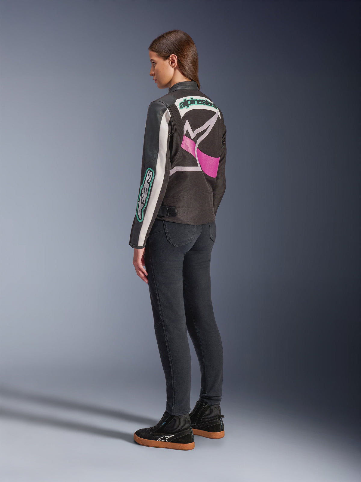 Alpinestars Idles Women’s Jacket, casaco de motociclismo, preto, off-white e roxo, vista de costas numa modelo, com um grande logótipo Alpinestars envolvente e design de inspiração vintage racing, combinado com calças de ganga escuras e sapatos pretos