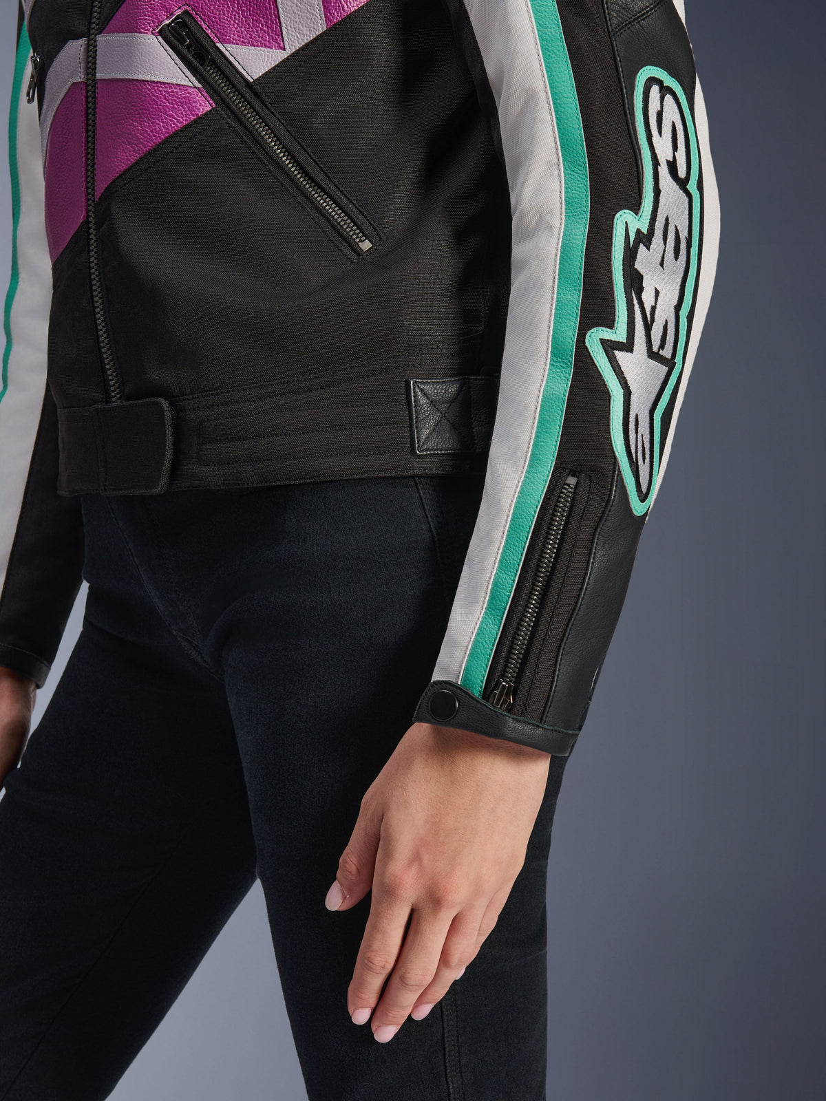 Casaco Feminino Alpinestars Idles, vista de pormenor da manga e da lateral do tronco, preto com detalhes em roxo, branco e verde-azulado, apresentando um logótipo Alpinestars marcante no braço, punho com fecho de correr e mola de pressão, e correia de ajuste na cintura, equipamento de motociclismo de inspiração vintage.