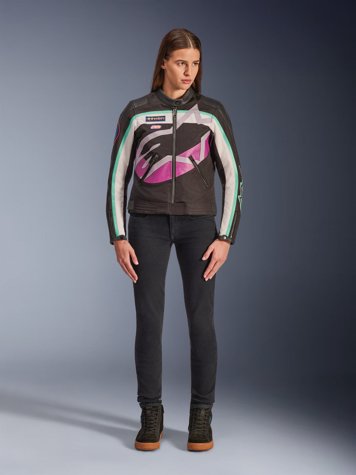 Mulher a usar o Casaco Feminino Alpinestars Idles, preto, off-white e roxo, design de inspiração vintage com logótipo envolvente grande e detalhes em verde-azulado, combinado com calças de ganga pretas e sapatilhas de motociclismo, vestuário urbano de motociclismo