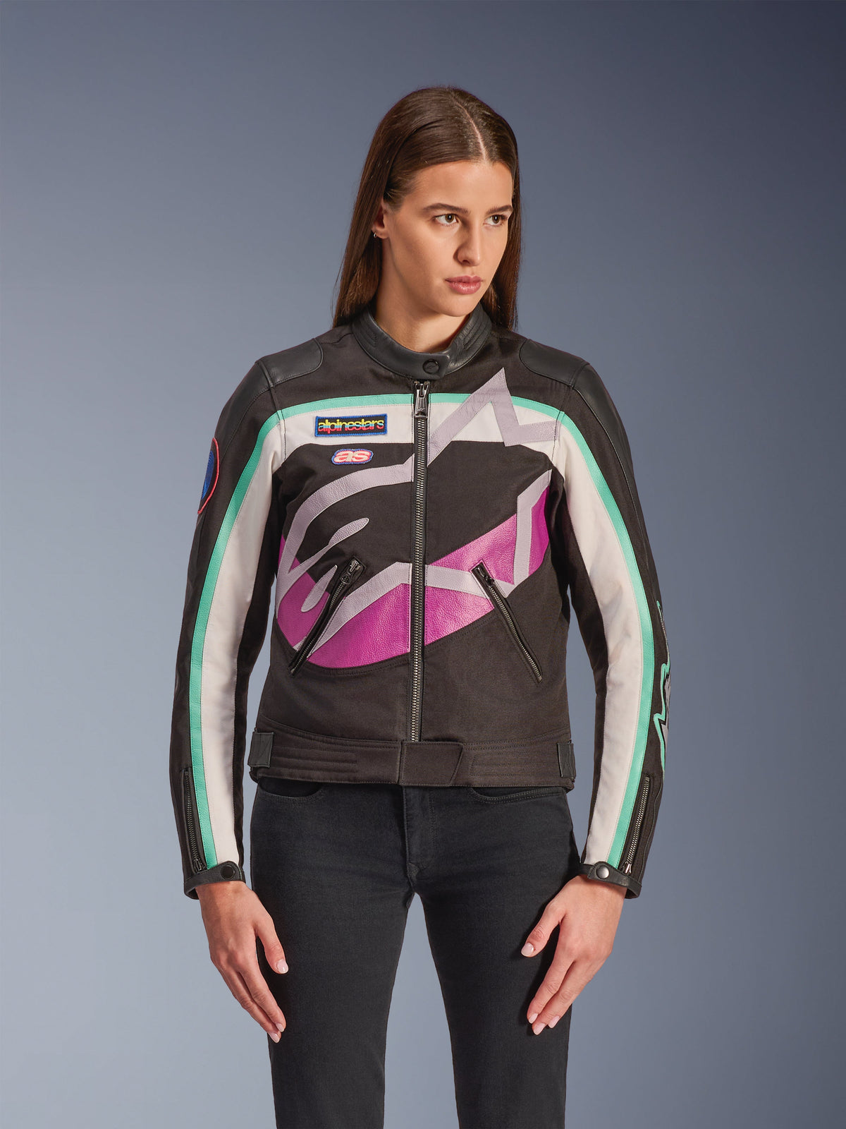 Casaco Alpinestars Idles para Mulher, casaco de motociclismo, preto com design color block em branco, roxo e verde-água, estilo de inspiração vintage racing com logótipo abstrato grande no peito, patches da marca e fecho frontal, usado por uma modelo com calças de ganga pretas