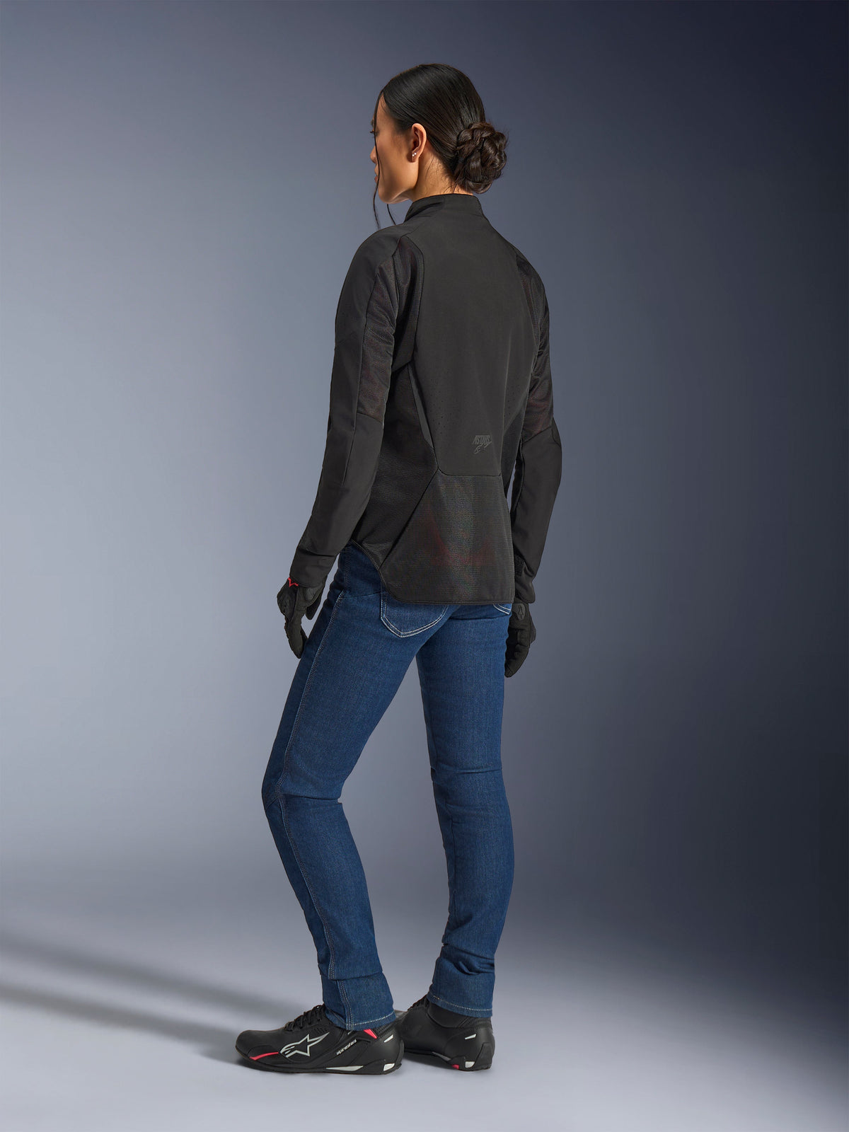 Woman wearing the Alpinestars Stella Aeroshell Airflow Jacket, Black Anthracite, rear three-quarter view showing breathable mesh panels and elongated rear profile, paired with blue denim riding jeans and black Alpinestars shoes