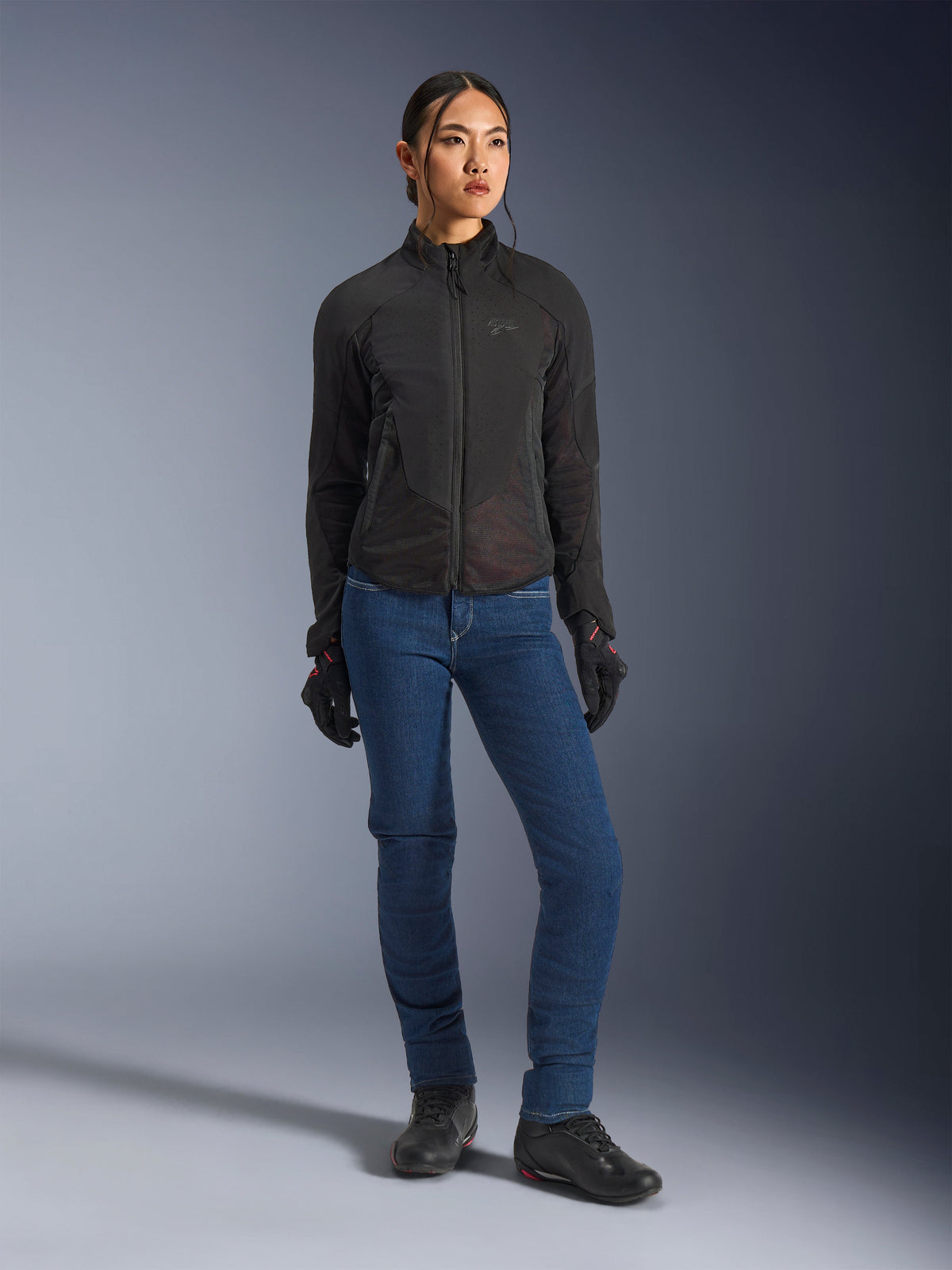 Female rider wearing the Alpinestars Stella Aeroshell Airflow Jacket in Black, featuring laser-perforated ventilation and mesh panels, styled with blue riding jeans and black motorcycle gear, modern sport and urban riding aesthetic