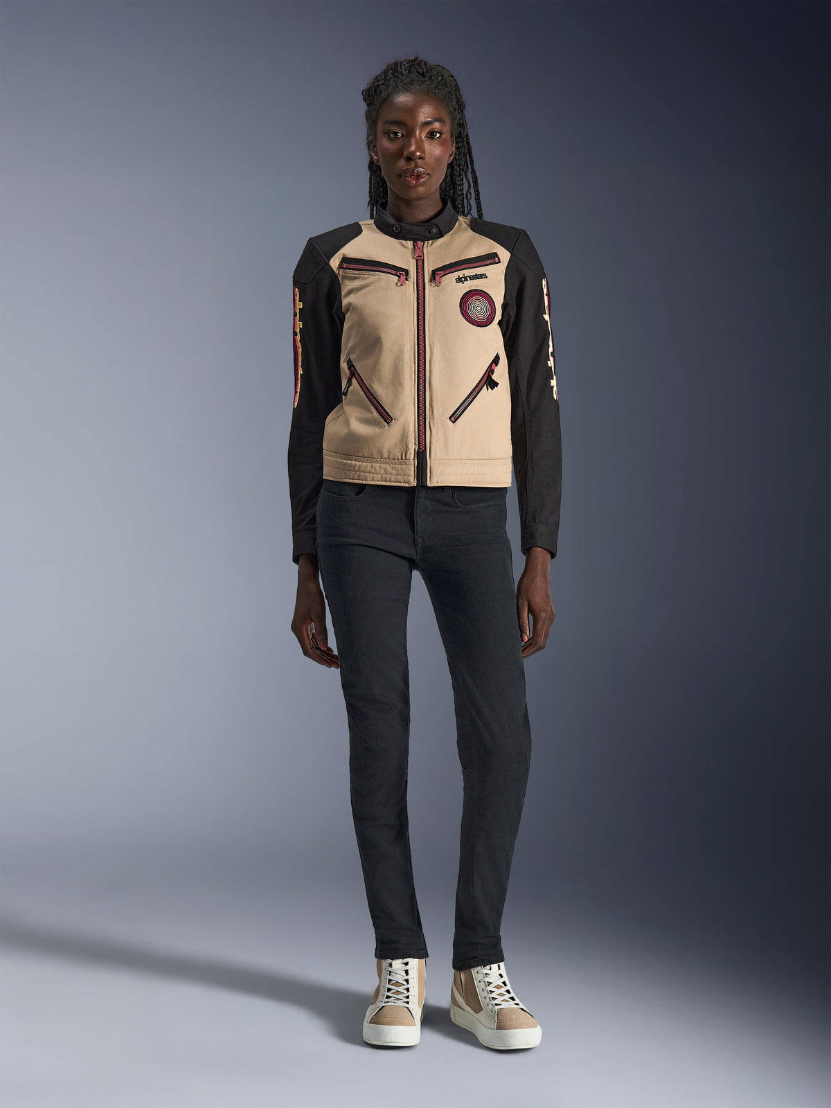 Casaco Stella Grace da Alpinestars para mulher, casaco de motociclismo urbano, construção têxtil em bege e preto, com fechos de correr frontais e patch bordado Oscar em espiral, usado com calças pretas e sapatilhas de cano alto, equipamento de condução contemporâneo inspirado em vestuário de trabalho