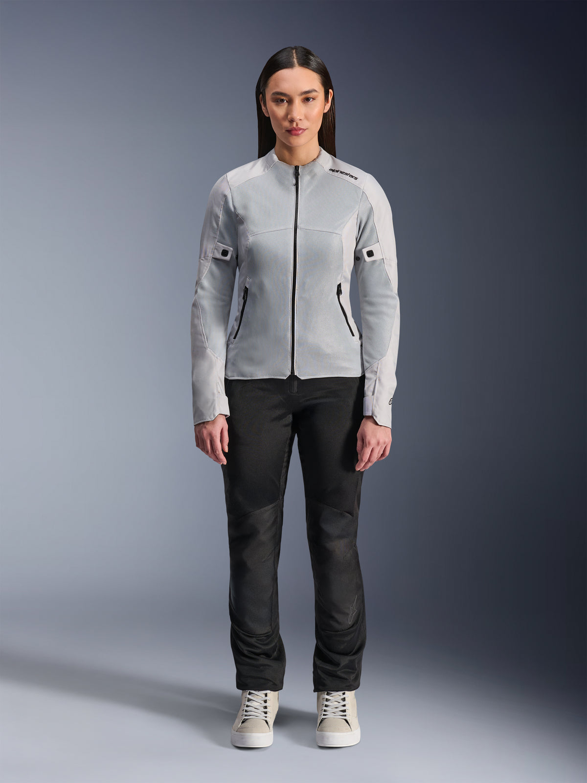 Mulher a usar o Casaco Alpinestars Stella C-1 Air em prateado, vista frontal, casaco têxtil de motociclismo urbano com painéis de malha técnica, combinado com calças pretas e sapatilhas brancas, equipamento de condução contemporâneo