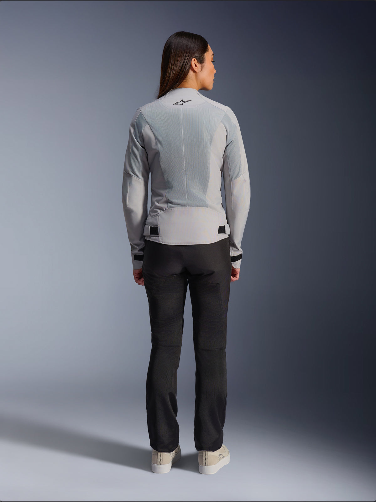Mulher a usar o Casaco Stella C-1 Air da Alpinestars em prateado, vista traseira, com grandes painéis de malha técnica para ventilação e logótipo de estrela refletor, calças de condução pretas e ténis brancos, equipamento de motociclismo urbano contemporâneo
